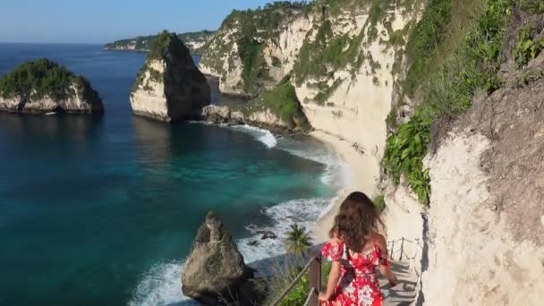 Brunette vrouw in een rode lange jurk gaat naar beneden op de trap tussen de rotsen en genieten van uitzicht op het paradijs het Diamond Beach in Nusa Penida eiland, Bali, Indonesië — Stockvideo