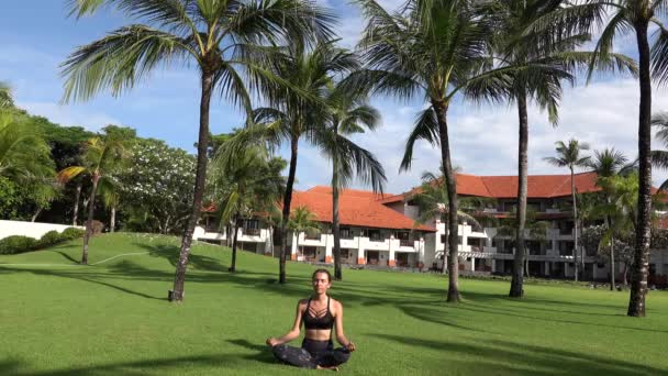 Sportig flicka sitter i lotus poserar gör yoga namaste munda händer poserar på grönt gräs i park med palmer. Yoga kvinna utomhus. Friska livsstil koncept — Stockvideo