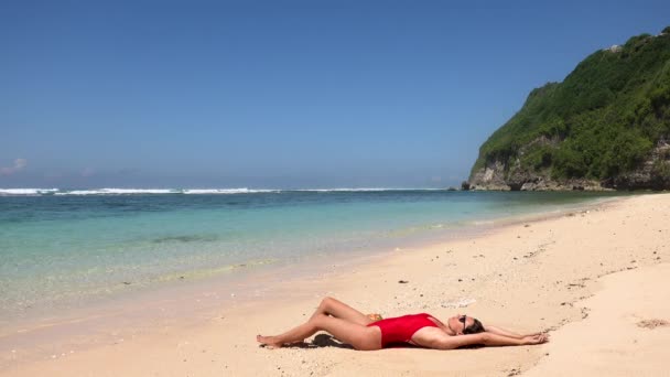 野生の熱帯砂のビーチの水の近くに横たわっている赤い水着の若い女性モデルとココナッツジュースを飲んで、バリ、インドネシア。旅行のコンセプトをリラックス — ストック動画