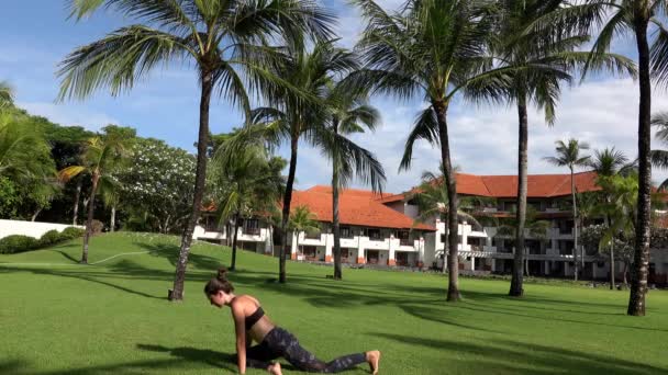 Yoga yapan genç sporcu kadın, yüzükoyun parkta palmiye ağaçlarıyla poz veriyor. Açık havada yoga yapan esnek bir kız. Genç bayan tropik arka planda yoga yapıyor. — Stok video