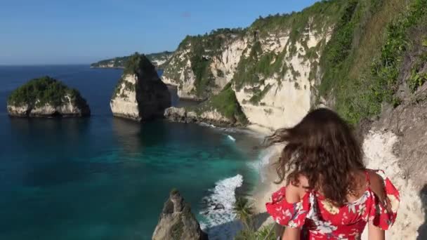 Πίσω όψη της μελαχρινής γυναίκας σε ένα κόκκινο φόρεμα πηγαίνει κάτω στις σκάλες μεταξύ των βράχων στο Diamond Beach στο νησί Nusa Penida, Μπαλί, Ινδονησία. Ταξιδιωτική φύση τροπική παραλία έννοια — Αρχείο Βίντεο