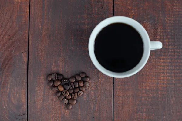 Форма сердца из кофейных зерен и чашка кофе на коричневом деревянном столе — стоковое фото