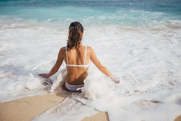 Back view of young woman in swimsuit sitting at white sand beach — Stock Photo, Image