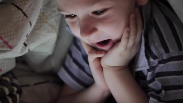 Мальчик лежит под одеялом и смотрит мультфильм на сенсорной панели — стоковое видео