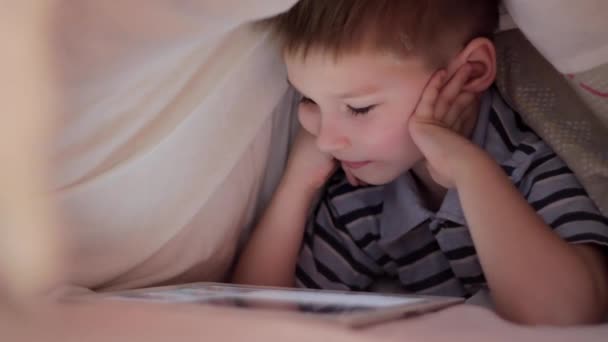 Niño acostado debajo de la manta y viendo dibujos animados en la almohadilla táctil — Vídeos de Stock