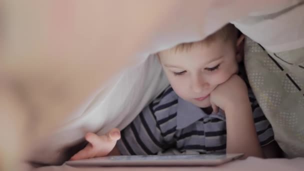 Мальчик лежит под одеялом и смотрит мультфильм на сенсорной панели — стоковое видео