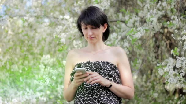 Genç kadın ile smartphone parkta. Bahar. Bir çiçekli ağaç arka planı — Stok video