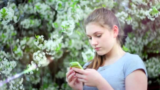 Chica adolescente con teléfono inteligente en el parque. Primavera. Sobre el fondo de un árbol floreciente — Vídeo de stock