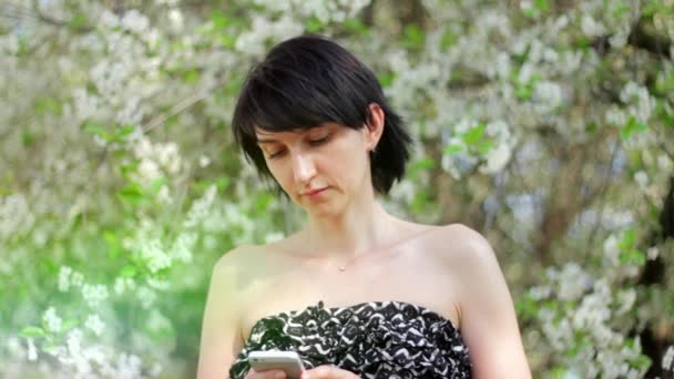 Jonge vrouw met smartphone in het park. Lente. Tegen de achtergrond van een bloeiende boom — Stockvideo