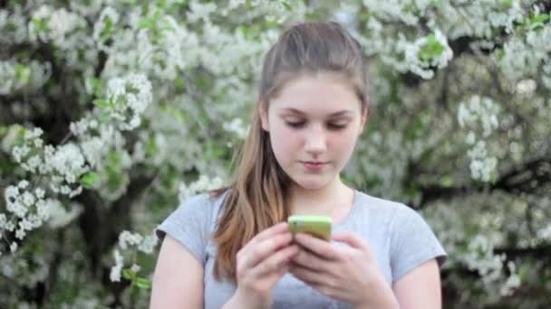 Chica adolescente con teléfono inteligente en el parque. Primavera. Sobre el fondo de un árbol floreciente — Vídeos de Stock