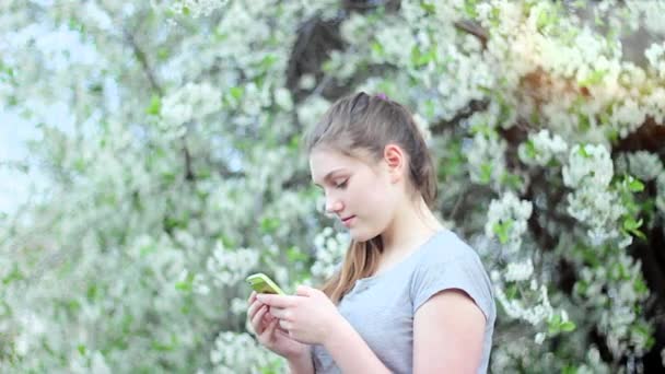 Teen flicka med smartphone i parken. Våren. Mot bakgrund av ett blommande träd — Stockvideo