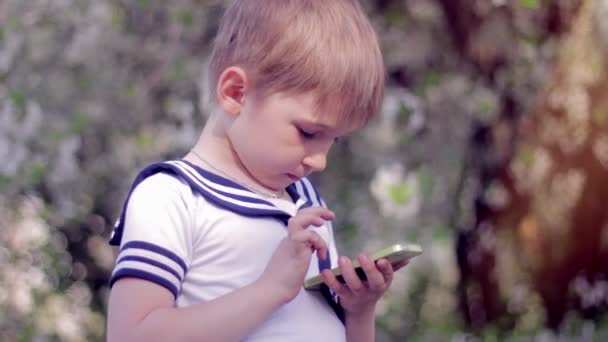 Young little boy plays games on smartphone. Spring park — Stock Video