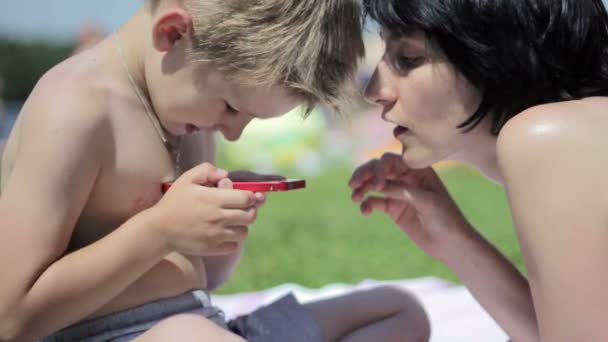 Niño pequeño con su madre en una playa jugando con un smartphone — Vídeos de Stock