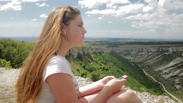 Adolescenta asculta muzica cu casti pe smartphone pe un fundal de munti — Videoclip de stoc