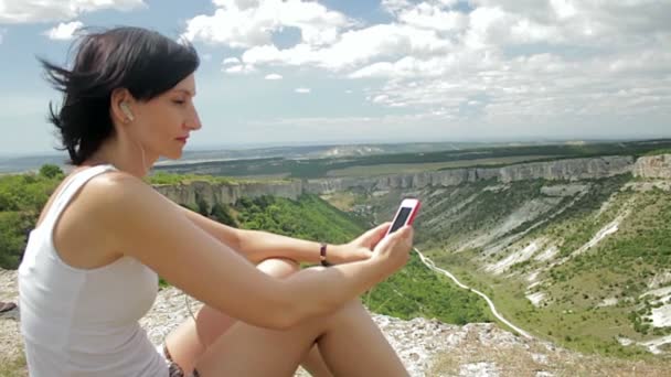 Приваблива жінка слухає музику з навушниками на смартфоні на фоні гір — стокове відео