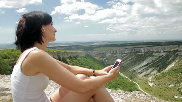 Donna attraente ascoltare musica con le cuffie su smartphone su uno sfondo di montagne — Video Stock