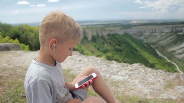 Kid boy listening to music with headphones on smartphone on a background of mountains — Stock Video