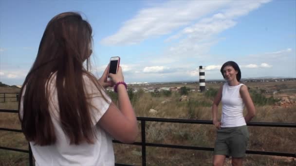 Счастливые женщины фотографируют друга со смартфоном на фоне морского маяка — стоковое видео