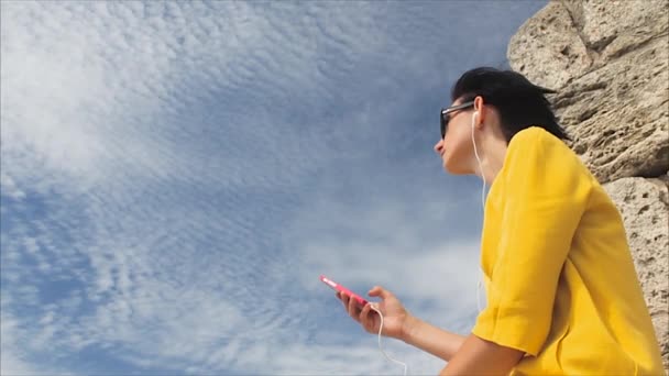 Mujer atractiva escuchando música con auriculares en el teléfono inteligente en un fondo del cielo y las ruinas de la antigua ciudad — Vídeo de stock