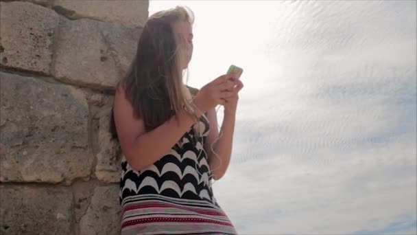 Chica adolescente escuchando música con auriculares en el teléfono inteligente en un fondo de mar y las ruinas de la antigua ciudad — Vídeo de stock