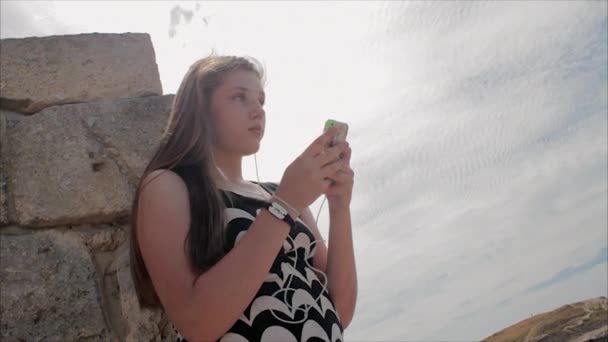 Teen girl listening to music with headphones on smartphone on a background of sea and the ruins of the ancient city — Stock Video