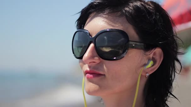 Close up portrait of beautiful young woman with headphones and sunglasses listening music on sea background — Stock Video
