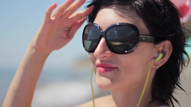 Close up portrait of beautiful young woman with headphones and sunglasses listening music on sea background — Stock Video