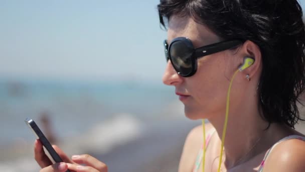 Крупный план портрета красивой молодой женщины в наушниках и солнцезащитных очках, слушающей музыку на смартфоне на море — стоковое видео