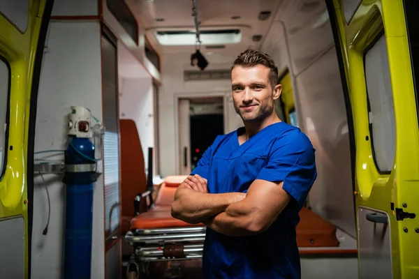 Hombre con uniforme médico mirando a la cámara y sonriendo, coche ambulancia en el fondo —  Fotos de Stock