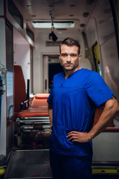 Hombre guapo con uniforme médico mirando a la cámara, coche ambulancia en el fondo —  Fotos de Stock