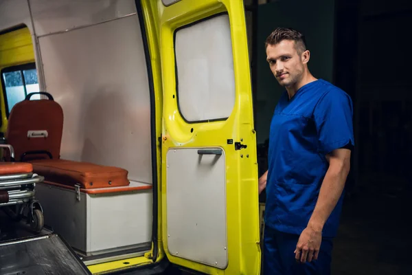 Un forense con uniforme azul se para en la parte trasera de un coche de ambulancia y mira a la cámara. —  Fotos de Stock