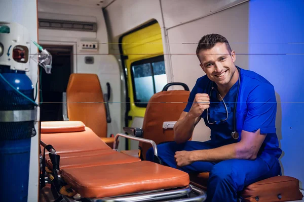 Joven hombre positivo en un uniforme médico sentado en el coche de la ambulancia y sonriendo con una sonrisa dentada —  Fotos de Stock