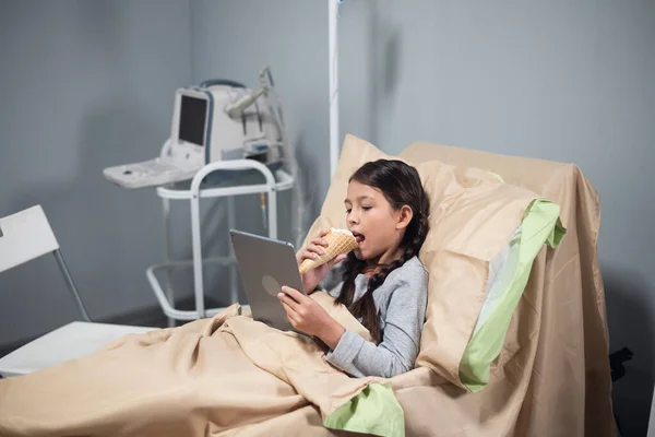 Niña morena comiendo un helado y viendo dibujos animados en su tableta en una cama de hospital — Foto de Stock