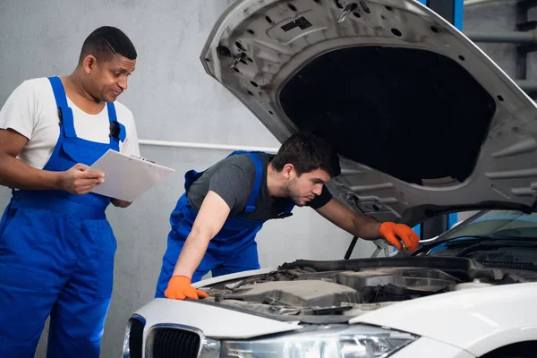 Mężczyźni w ubraniach roboczych naprawić silnik samochodu i napisać raport — Zdjęcie stockowe