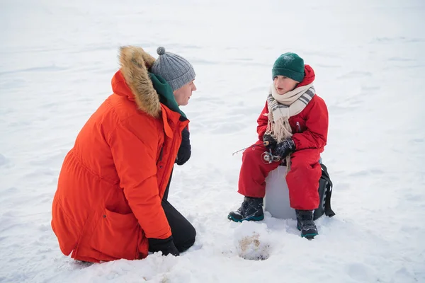 Un niño pequeño en un mono rojo y bufanda está pescando en un agujero de hielo con su padre —  Fotos de Stock