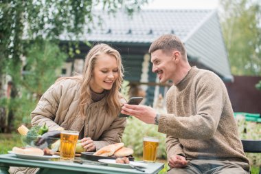 Tecrit edilmiş bir çift akıllı telefonlarından ilginç video bloglarını izleyerek barbekü yapıp rahatlıyor. Önlerinde abur cubur ve bira dolu bir masa var..