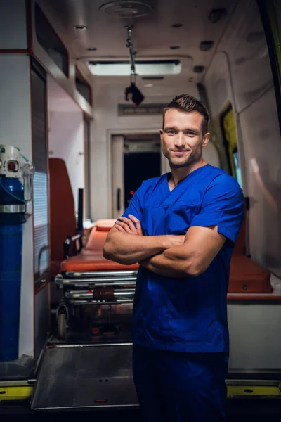 Un forense sonriente con uniforme médico se para con un coche de ambulancia en el fondo —  Fotos de Stock