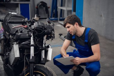 İşçi motosikleti kontrol eder ve notlar yazar.