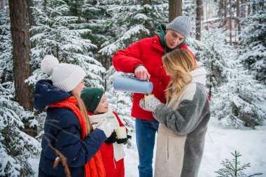 İki çocuklu aktif bir aile, şehrin dışındaki kış kar ormanlarında gezer ve sıcak tutmak için termostan sıcak kahve içer..