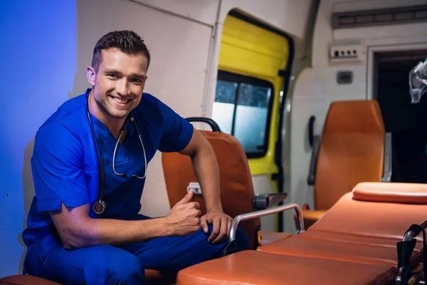 Un retrato de un joven paramédico sonriente con un uniforme azul sentado en el coche de la ambulancia y mostrando los pulgares hacia arriba —  Fotos de Stock