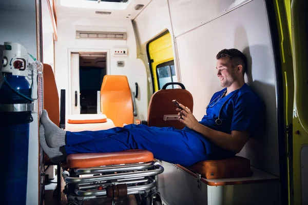 Мужчина парамедик в синей форме отдыхает и просматривает свой телефон в машине скорой помощи — стоковое фото