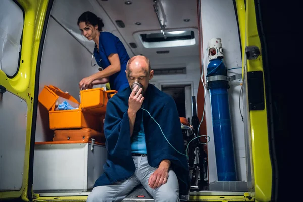 Man rescued from the fire sitting in an ambulance car, breathing through an oxygen mask, a female paramedic in the background — Stock fotografie