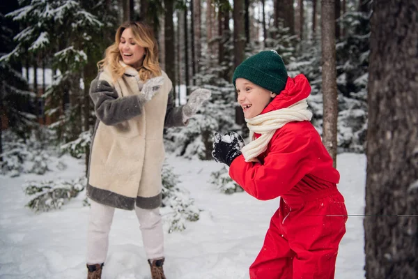 Filho em macacões de inverno e mãe em casaco de pele jogar neve na floresta no inverno — Fotografia de Stock