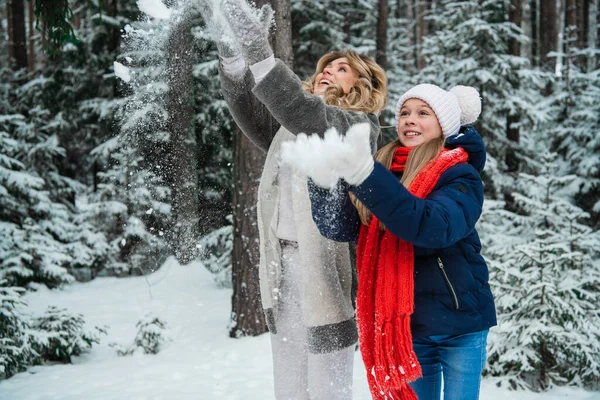 Uma jovem mulher bonita em um casaco de pele e uma menina bonita em uma jaqueta, chapéu e cachecol vomitar neve no inverno na floresta. — Fotografia de Stock