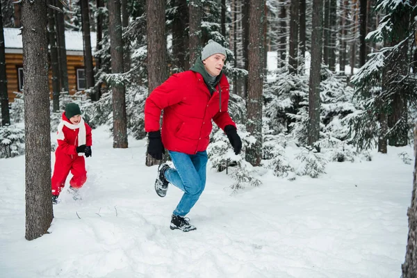 Мужчина в красной куртке в зимнем лесу, играет в догонялки со своим племянником, мальчик в красном зимнем комбинезоне и шарфе — стоковое фото