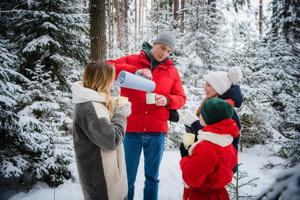 Anne ve baba çocuklarla birlikte karla kaplı bir ormanda çam ağaçlarıyla kışın tadını çıkarmak için dışarı çıktılar. Termosta lezzetli bir sıcak çay..