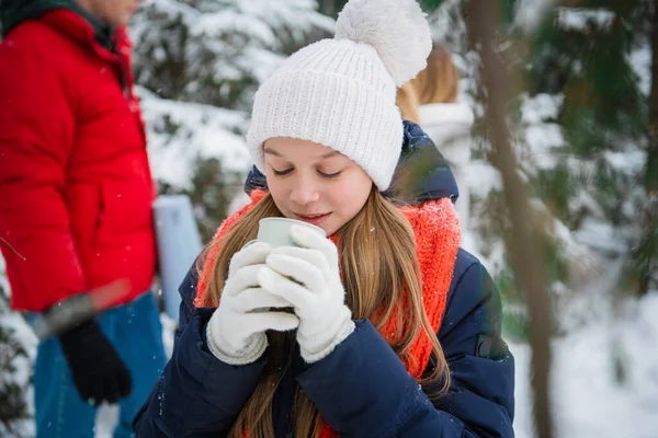 A young long-haired blonde girl drinks tea from a mug in the winter forest. Portrait with depth of field against the background of fir trees and snowdrifts.