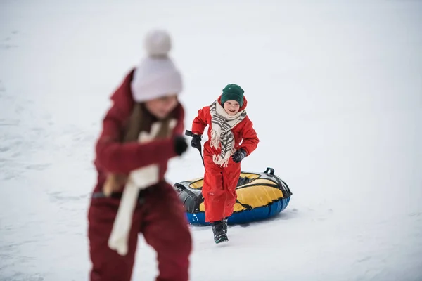 Χειμερινός περίπατος παιδιών με κόκκινες φόρμες, ένα αγόρι κουβαλάει τις σωληνώσεις του στο λόφο. — Φωτογραφία Αρχείου
