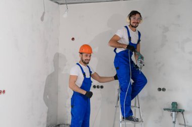 A builder in overalls stands on a stepladder, his colleague helps him