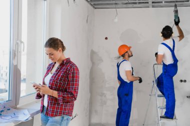 Man in overalls use drill and stepladder, woman typing on the phone